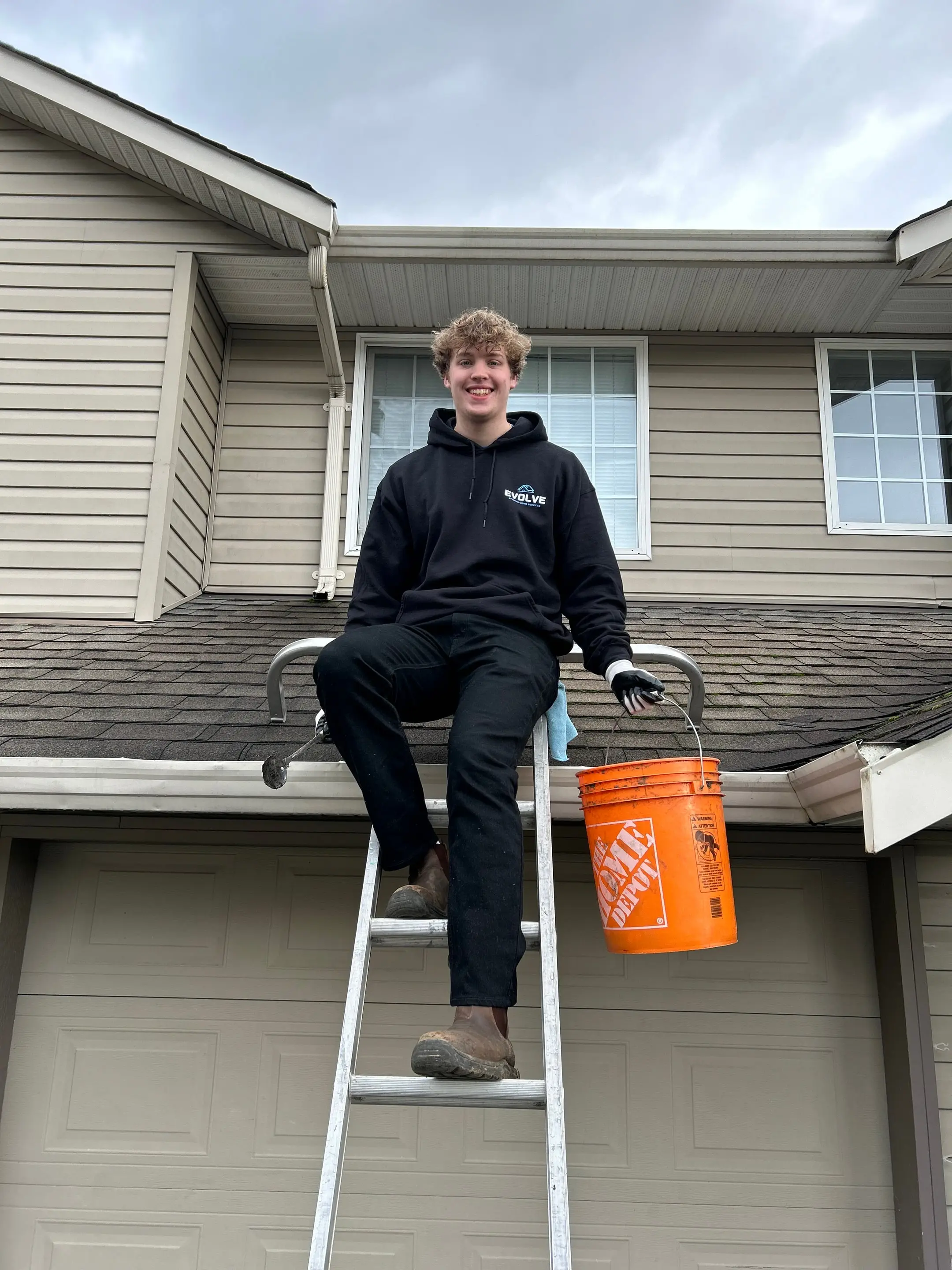 Smiling Evolve crew member on a ladder at the gutter line of a two-storey home, with an orange debris bucket