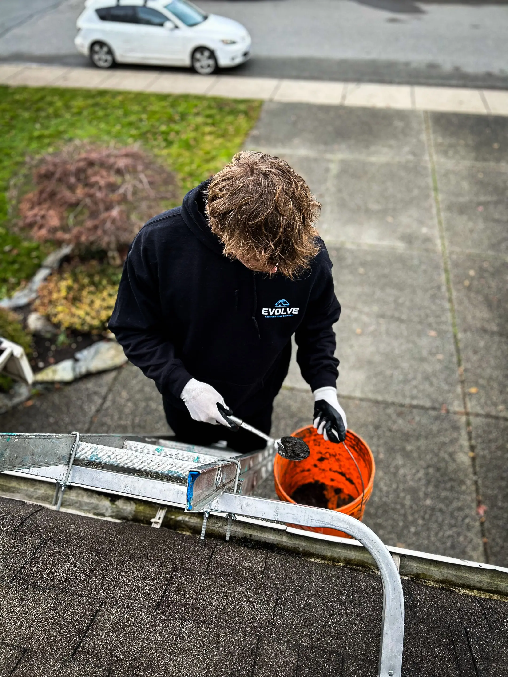 Crew member in an Evolve hoodie on a ladder scooping gutter sludge into an orange bucket