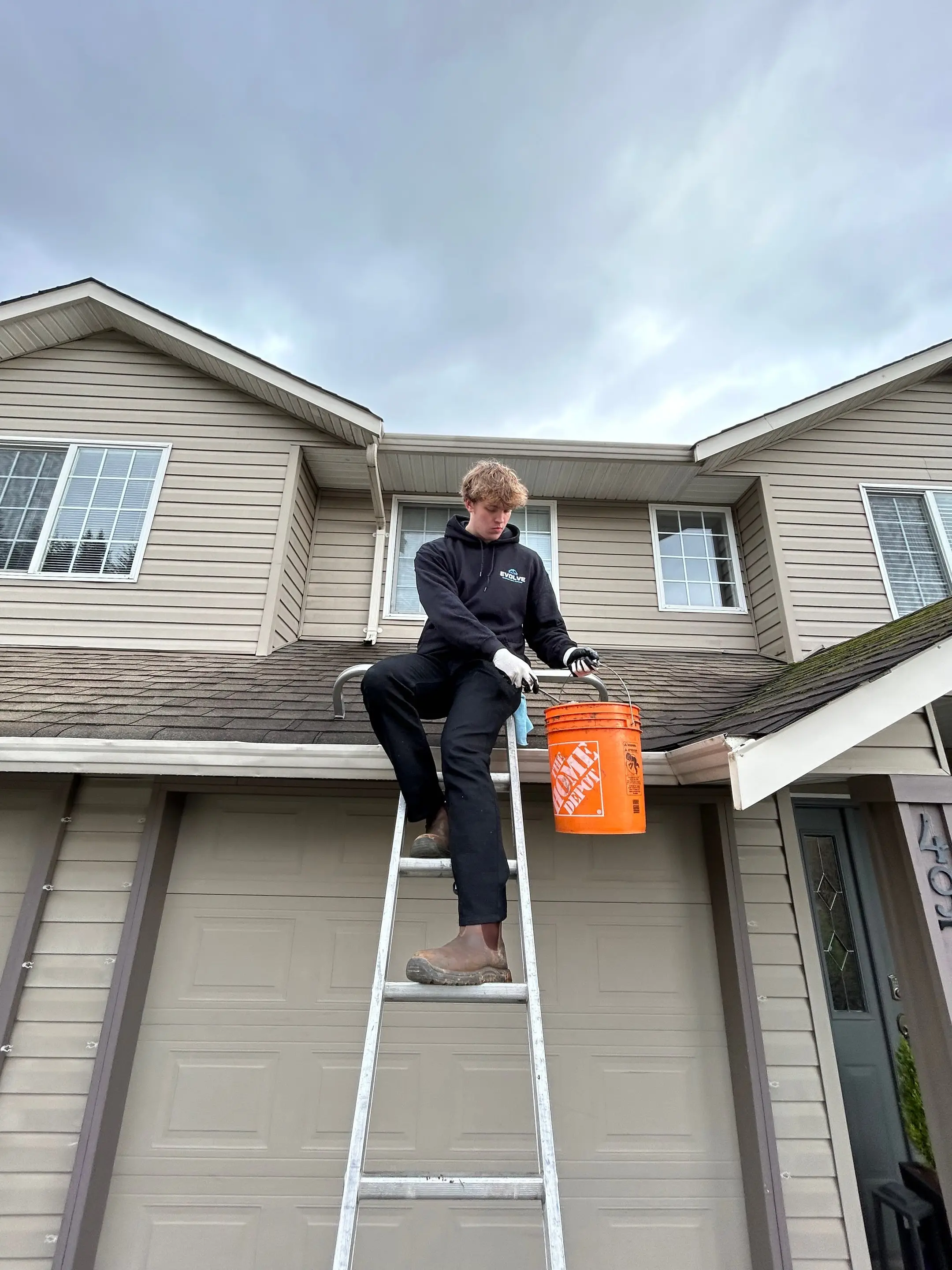 Evolve crew member in a hoodie on a ladder at the gutter line of a two-storey home, with an orange debris bucket