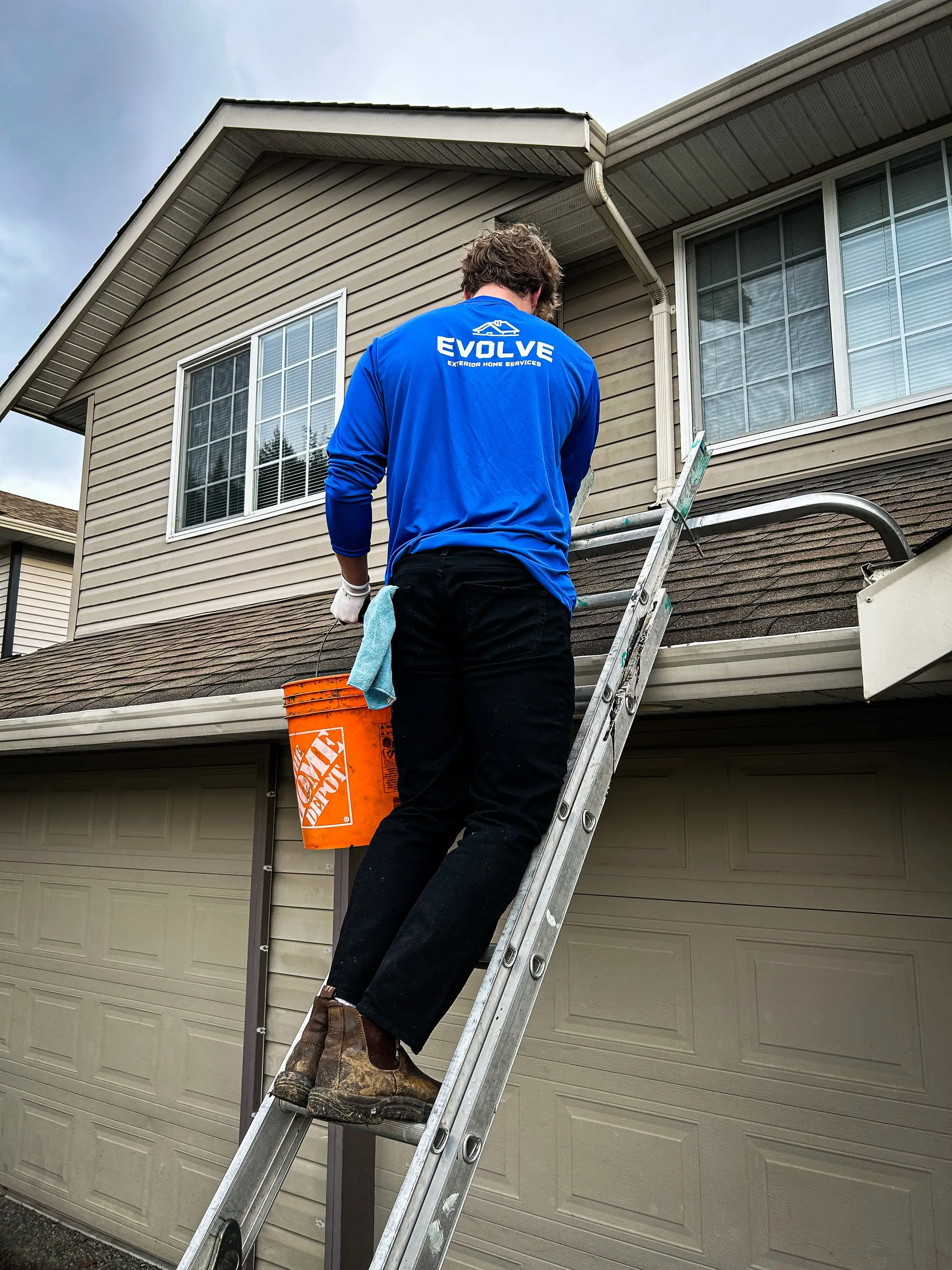 Evolve crew member on a ladder with an orange bucket, clearing the gutter above the garage of a two-storey home