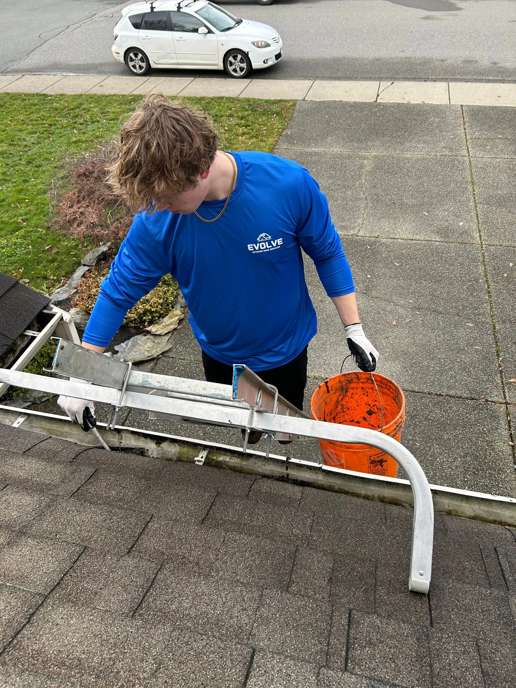 Evolve crew member at the roof edge scooping wet debris out of a gutter into an orange bucket