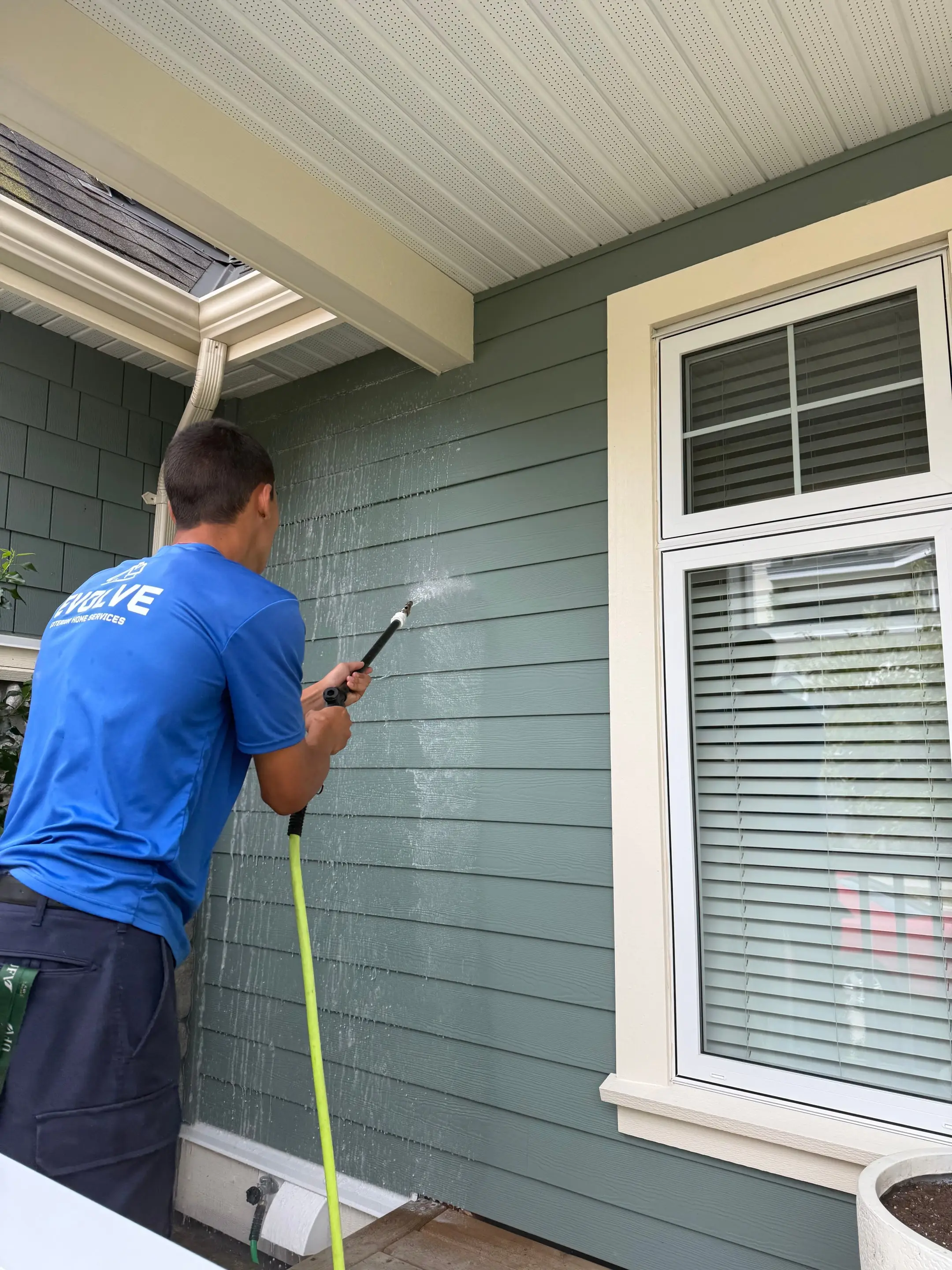 Evolve crew member softwashing green lap siding beside a front window with a low-pressure wand