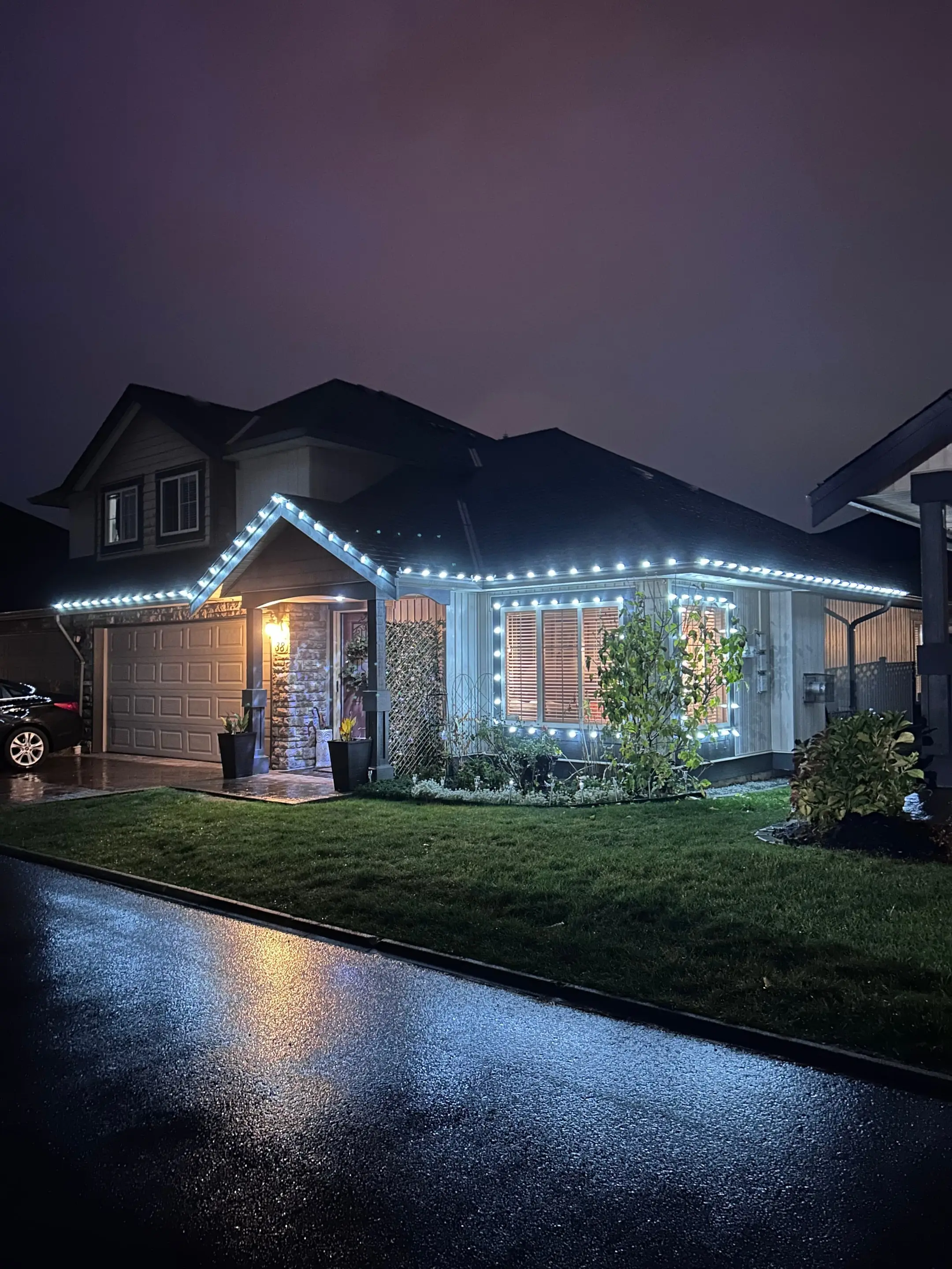 Grey two-storey home at night outlined in cool white Christmas lights, reflected on a wet street